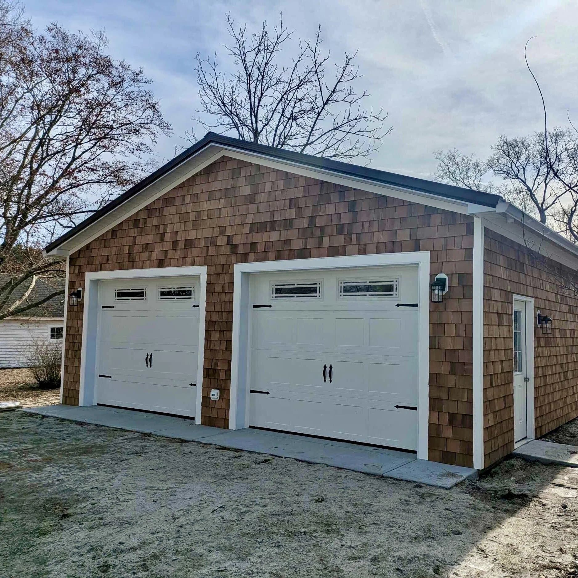Detached cedar-shake garage with two white carriage-style garage doors with black hardware