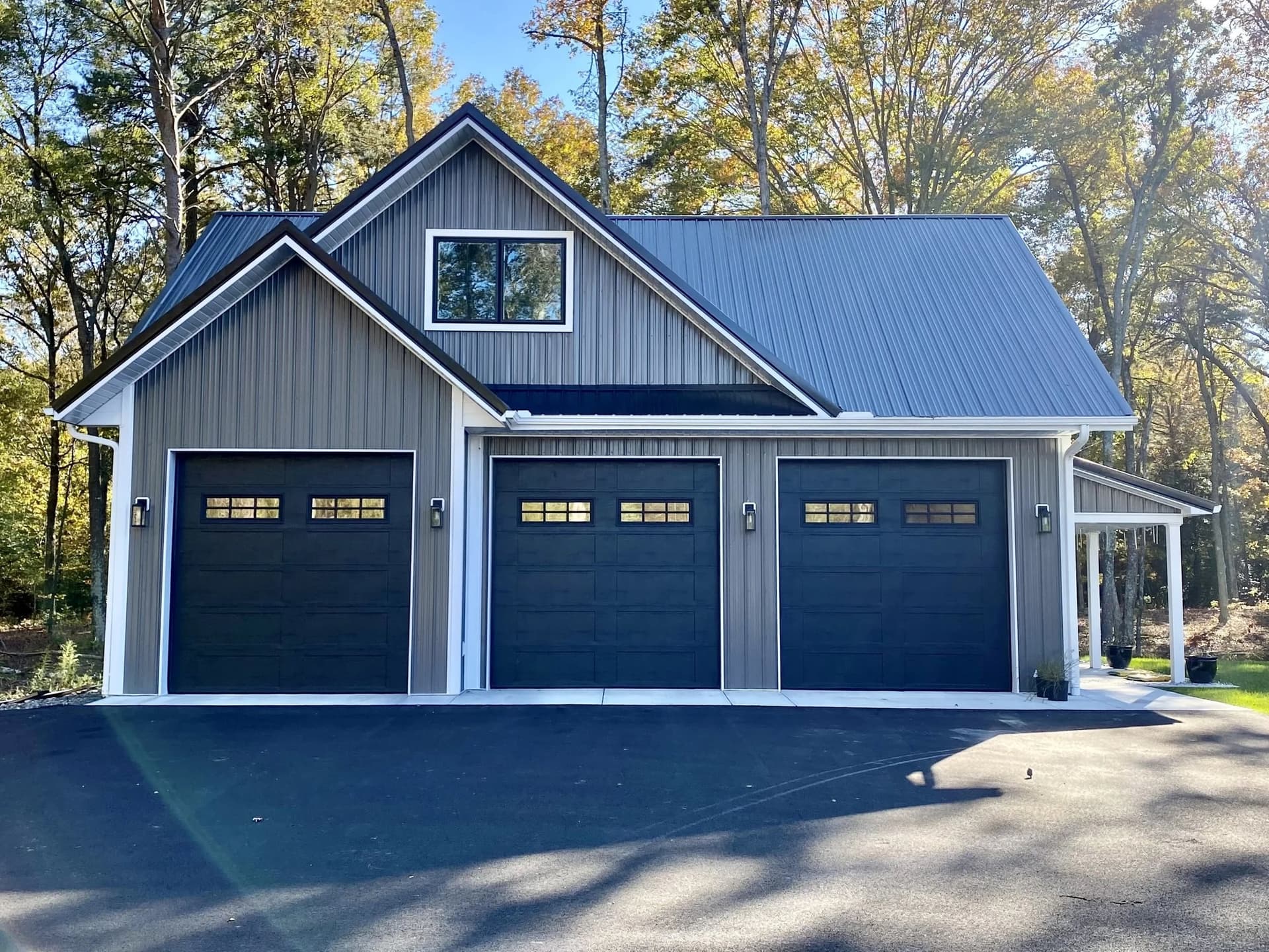 Gray home with a blue metal roof and three black garage doors with window tops, in the woods