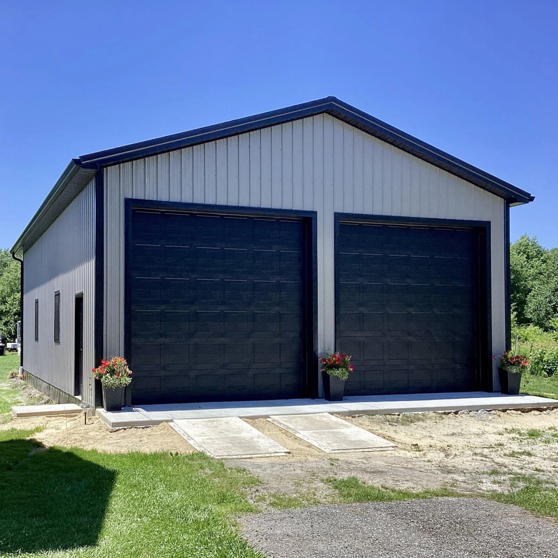 Gray metal building with two tall black garage doors and flower pots on a gravel drive