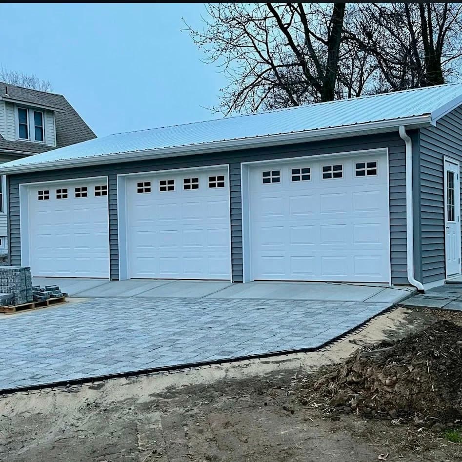 Three white raised-panel garage doors with window tops on a gray-sided three-car garage with a paver drive